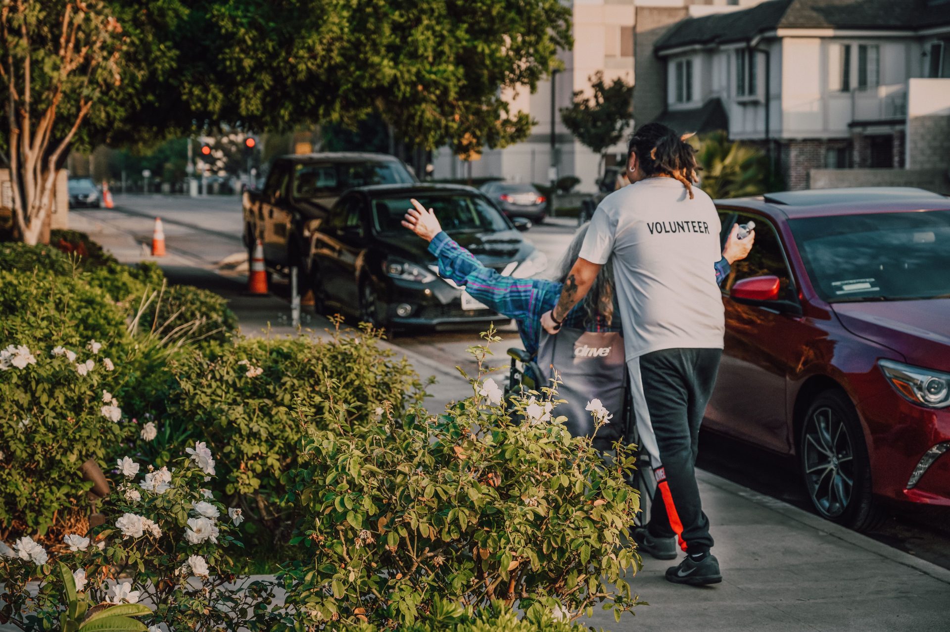 Volunteer assisting a person in a wheelchair on a city street surrounded by greenery and parked cars.