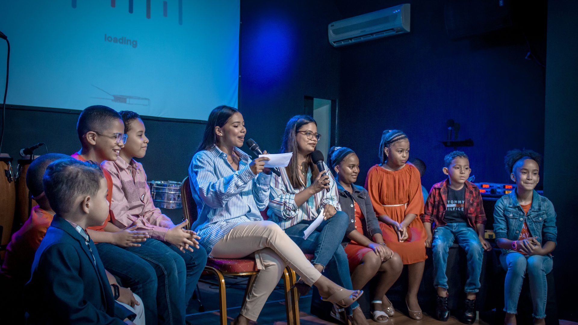 A group of children and young women participating in a panel discussion indoors.